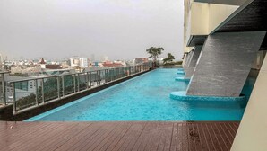 A rooftop swimming pool with a wooden deck, surrounded by a glass railing and a cityscape view. - Menteng Park By The Jones (Jakarta)