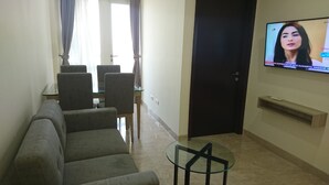 A living room with a sofa, a glass coffee table, a TV mounted on the wall, and a dining area with chairs and a table. - Menteng Park By The Jones (Jakarta)
