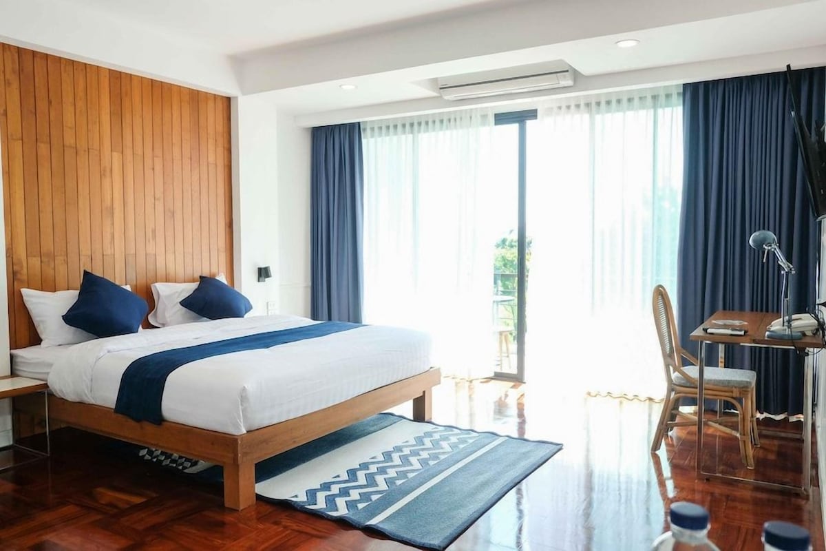 Une chambre d’hôtel avec une tête de lit en bois, un lit avec une literie blanche et des touches de bleu, un bureau avec une chaise et une grande fenêtre avec des rideaux.