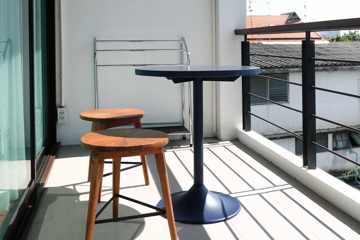 Een balkon met een tafel en twee krukken, een glazen deur en uitzicht op gebouwen.