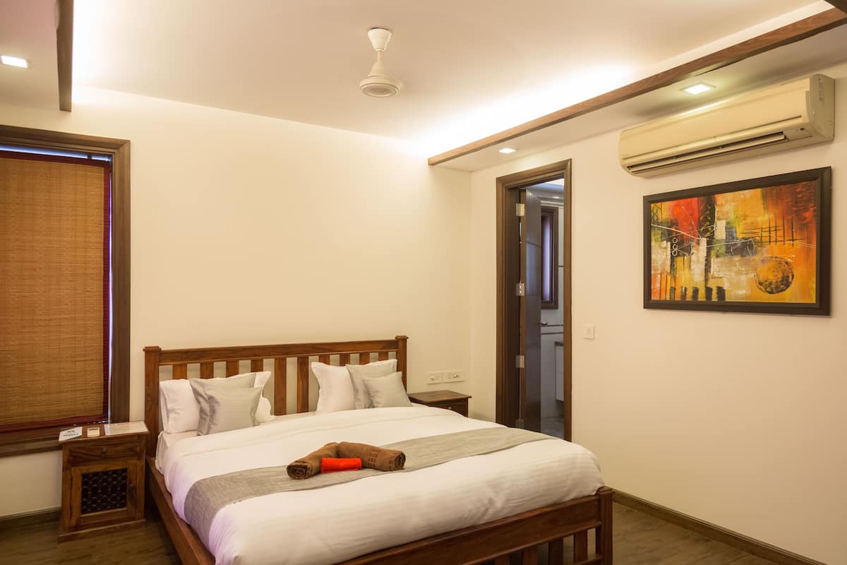 A hotel room with a wooden bed, a nightstand, an air conditioning unit, and a painting on the wall.