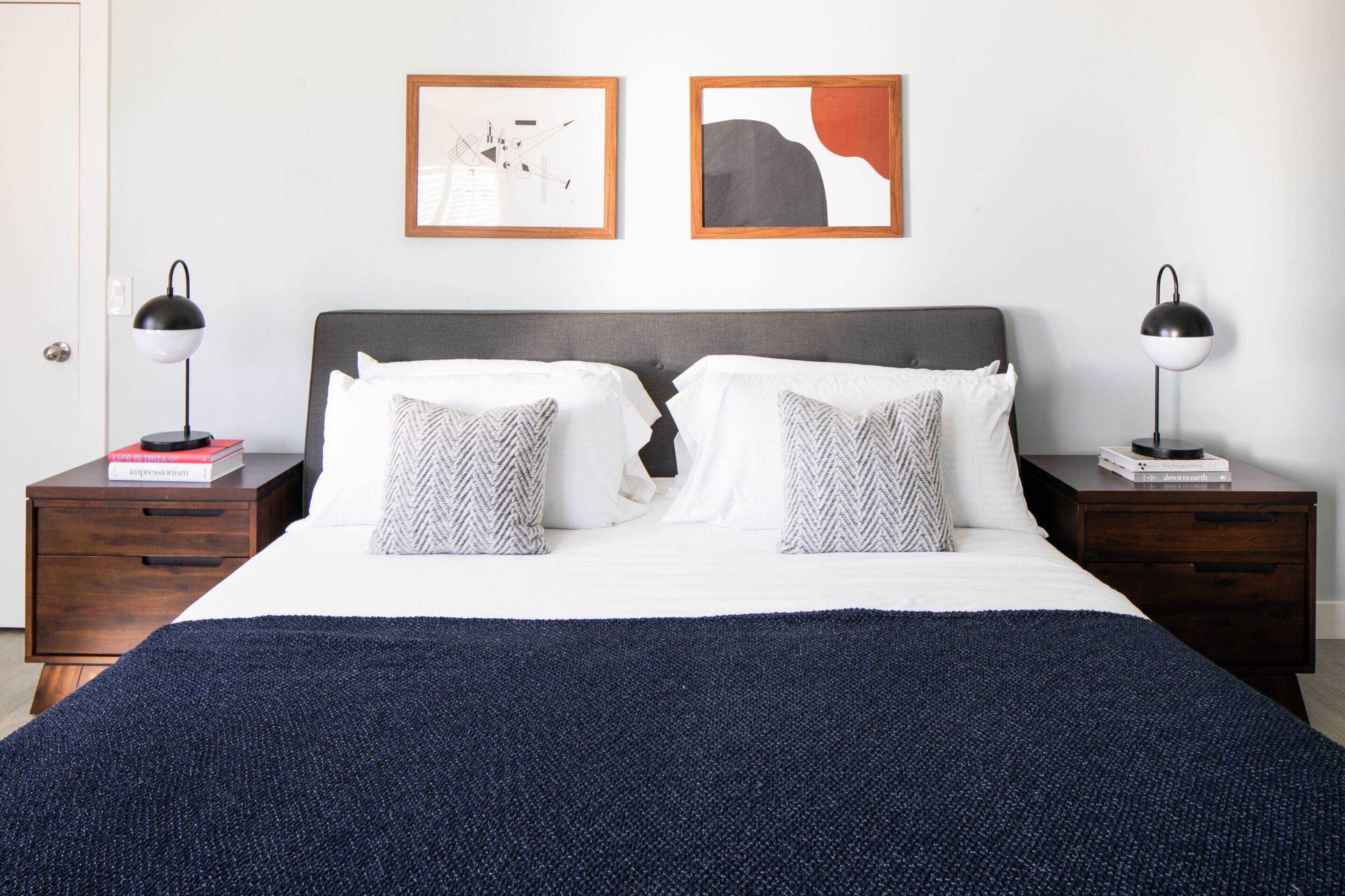 A double bed with a dark blue blanket, two bedside tables with lamps, and two framed artworks on the wall.