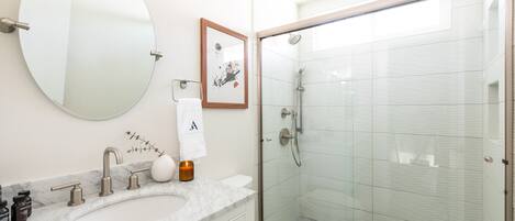 Un baño moderno con un espejo redondo, un lavabo de mármol, un cuadro enmarcado y una ducha con mampara de vidrio.