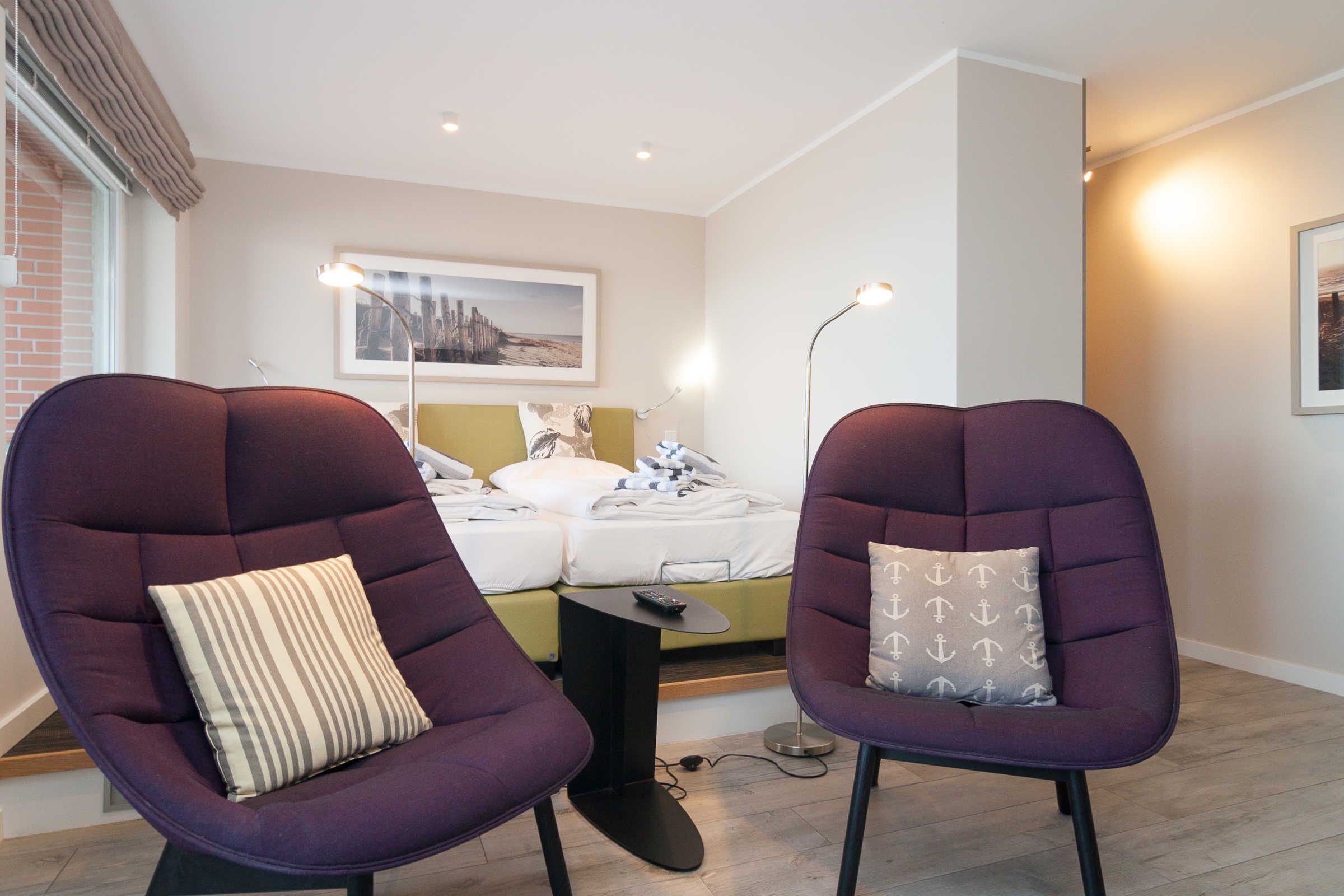 A modern living room with two purple armchairs, a sofa, a coffee table, and a bed with a headboard. There are framed pictures on the walls and a floor lamp.