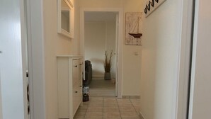 A narrow hallway with white walls and tiled flooring, leading to a room with a potted plant and a framed picture on the wall. - Budersand 26, Germany (Hörnum (Sylt))
