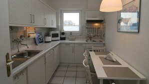 A compact kitchen with white cabinets, a built-in microwave, a sink, and a dining area with a table and chairs. - Budersand 26, Germany (Hörnum (Sylt))