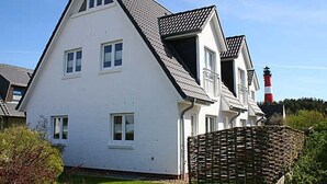 A white two-story house with a gabled roof, a visible chimney, and a red and white lighthouse in the background. - House Emilia app. lighthouse (Hörnum (Sylt))