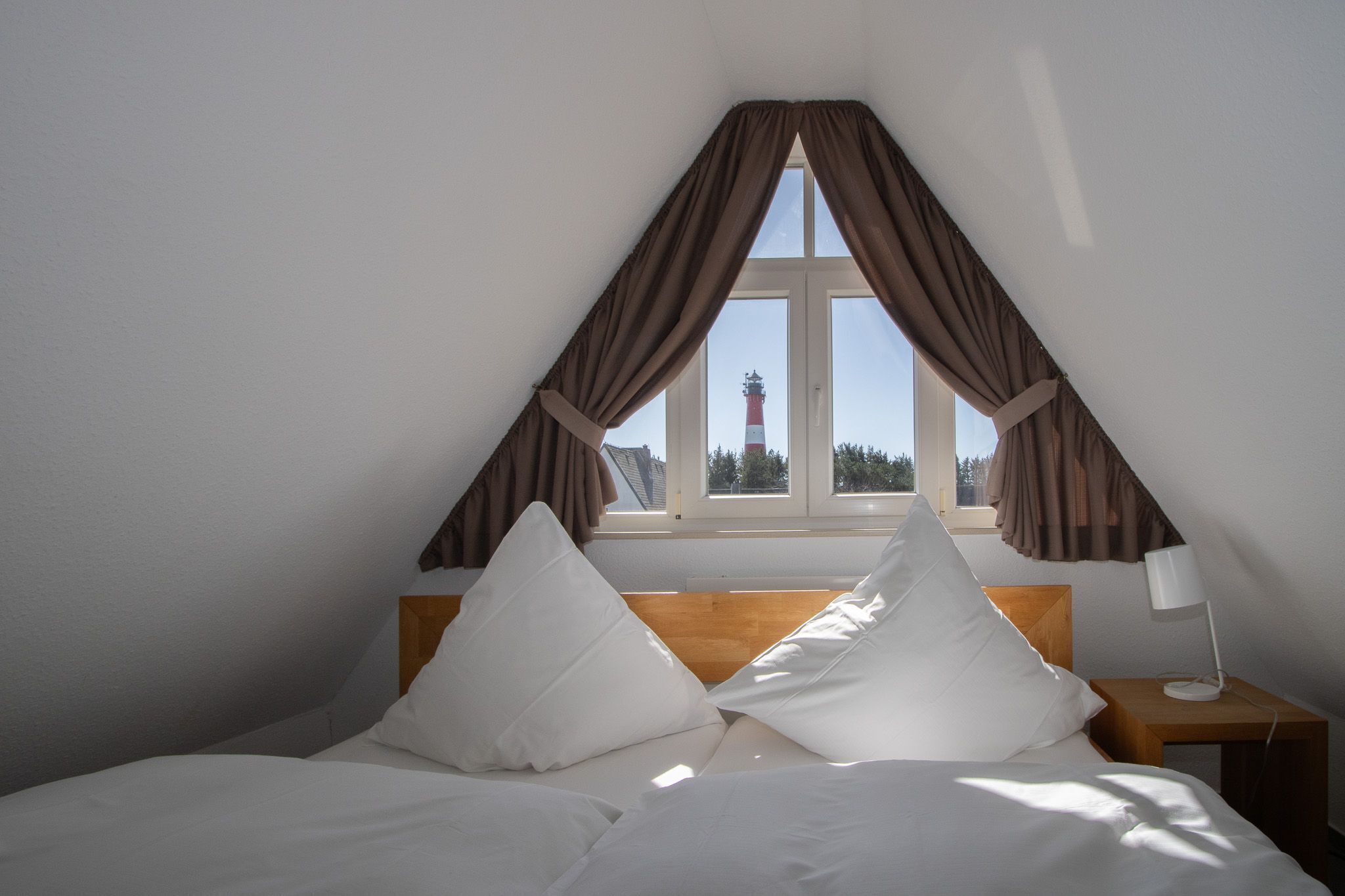 Une chambre à coucher avec une tête de lit en bois, deux oreillers blancs, une table de chevet avec une lampe et une fenêtre avec des rideaux.