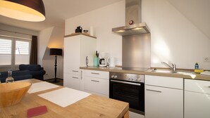 A modern kitchen with white cabinets, a built-in oven, a stainless steel range hood, and a wooden dining table with a red notebook. - House Emilia app. lighthouse (Hörnum (Sylt))