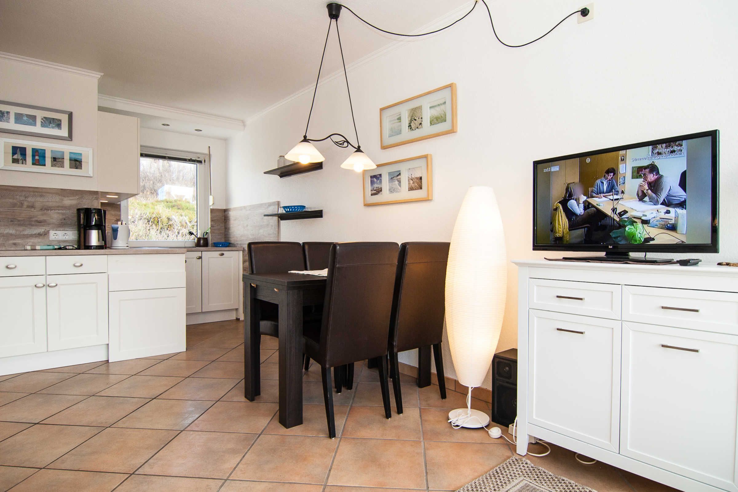 A modern kitchen with white cabinets, a dining table with chairs, a TV, and a pendant light.
