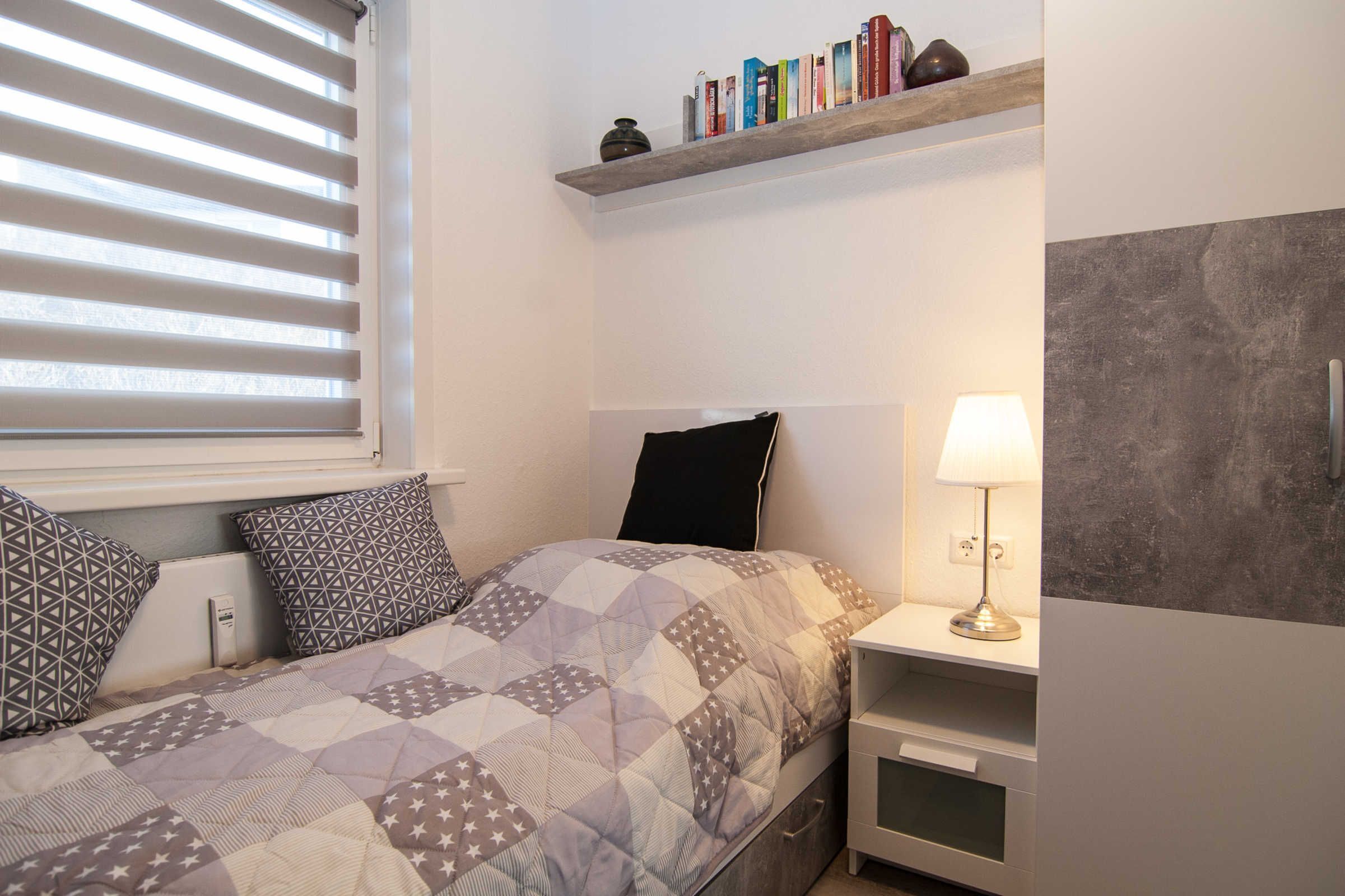 Une chambre à coucher avec un lit, des oreillers, une table de chevet, une lampe, une étagère avec des livres et une fenêtre avec des stores.
