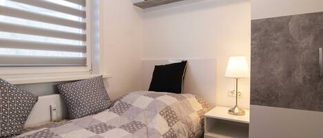 A bedroom with a bed, pillows, a nightstand, a lamp, a shelf with books, and a window with blinds.
