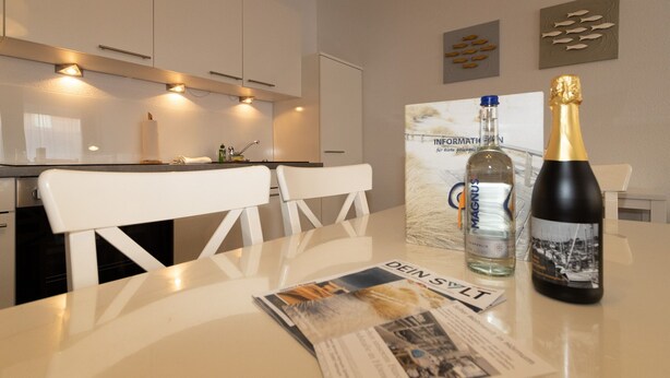 A modern kitchen with white cabinetry, a dining table with a magazine and a bottle of champagne. - Holiday beacon (Hörnum (Sylt))