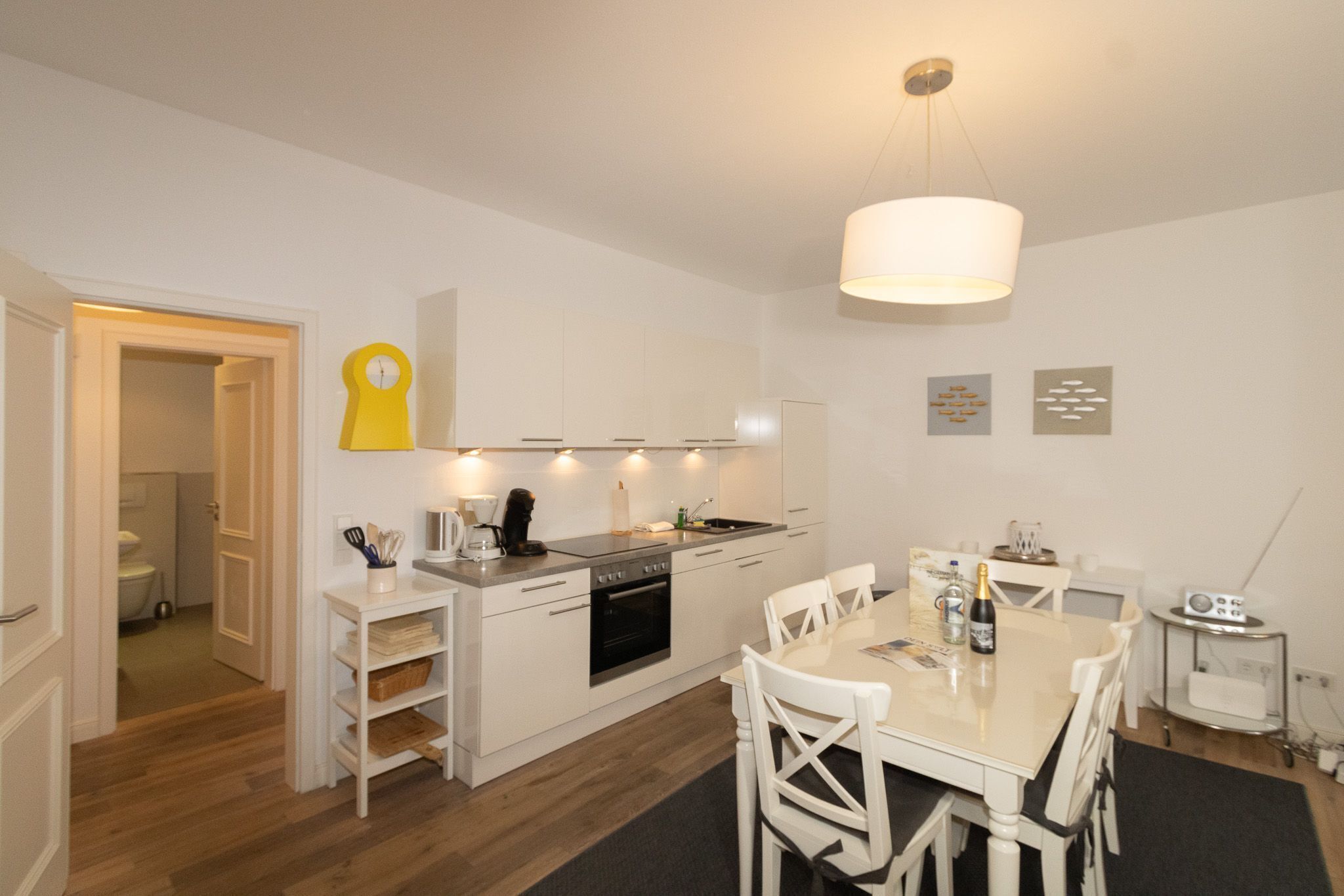 Een moderne keuken met witte kasten, een eettafel en stoelen. Er hangt een gele klok aan de muur en een radio op een bijzettafeltje.