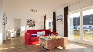 Ein geräumiges Wohnzimmer mit einem roten Sofa, einem Holztisch und Blick auf geparkte Autos durch große Fenster.