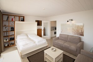 A bedroom with a bed, a sofa, a small table, a bookshelf, and a kitchen area.