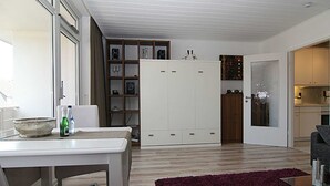 Moderni olohuone, jossa on ruokailutila, suuri valkoinen vaatekaappi ja avoimen oven kautta näkyvä keittiö.