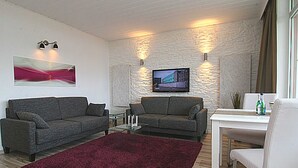 Olohuoneessa on kaksi sohvaa, sohvapöytä, seinälle asennettu televisio ja punainen matto. Seinän pinta on kuvioitu.