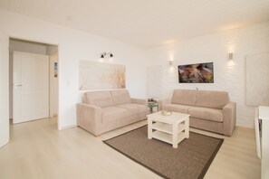 A living room with two beige sofas, a coffee table, and a rug. There are wall-mounted lights and a framed picture. - Holiday Ferlings (Hörnum (Sylt))