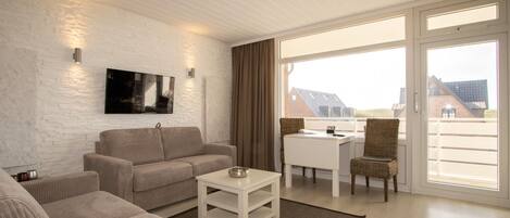 A modern living room with a sofa, a coffee table, and a TV mounted on a white brick wall. There is a balcony with a wicker chair and a table.