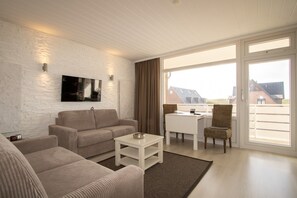 A modern living room with a sofa, a coffee table, and a TV mounted on a white brick wall. There is a balcony with a wicker chair and a table.