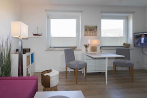 Ein modernes Wohnzimmer mit einem weißen Tisch, zwei grauen Stühlen, einem weißen Sofa und einem kleinen Holzschemel. Eine Fernseher befindet sich auf der rechten Seite.