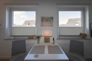 Ein Zimmer mit zwei Fenstern, einem Tisch mit Blumenvase und einem Gemälde an der Wand.