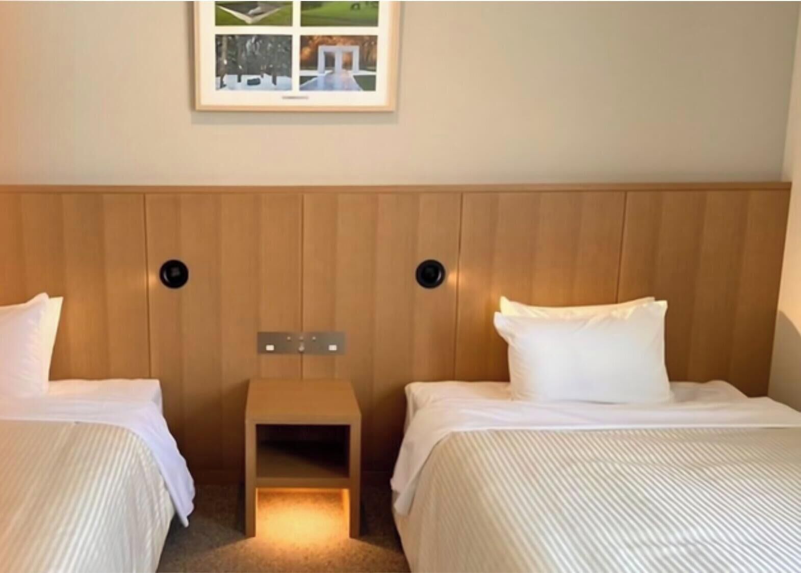 Een hotelkamer met twee bedden, een houten hoofdbord, een klein nachtkastje en een ingelijst schilderij aan de muur.