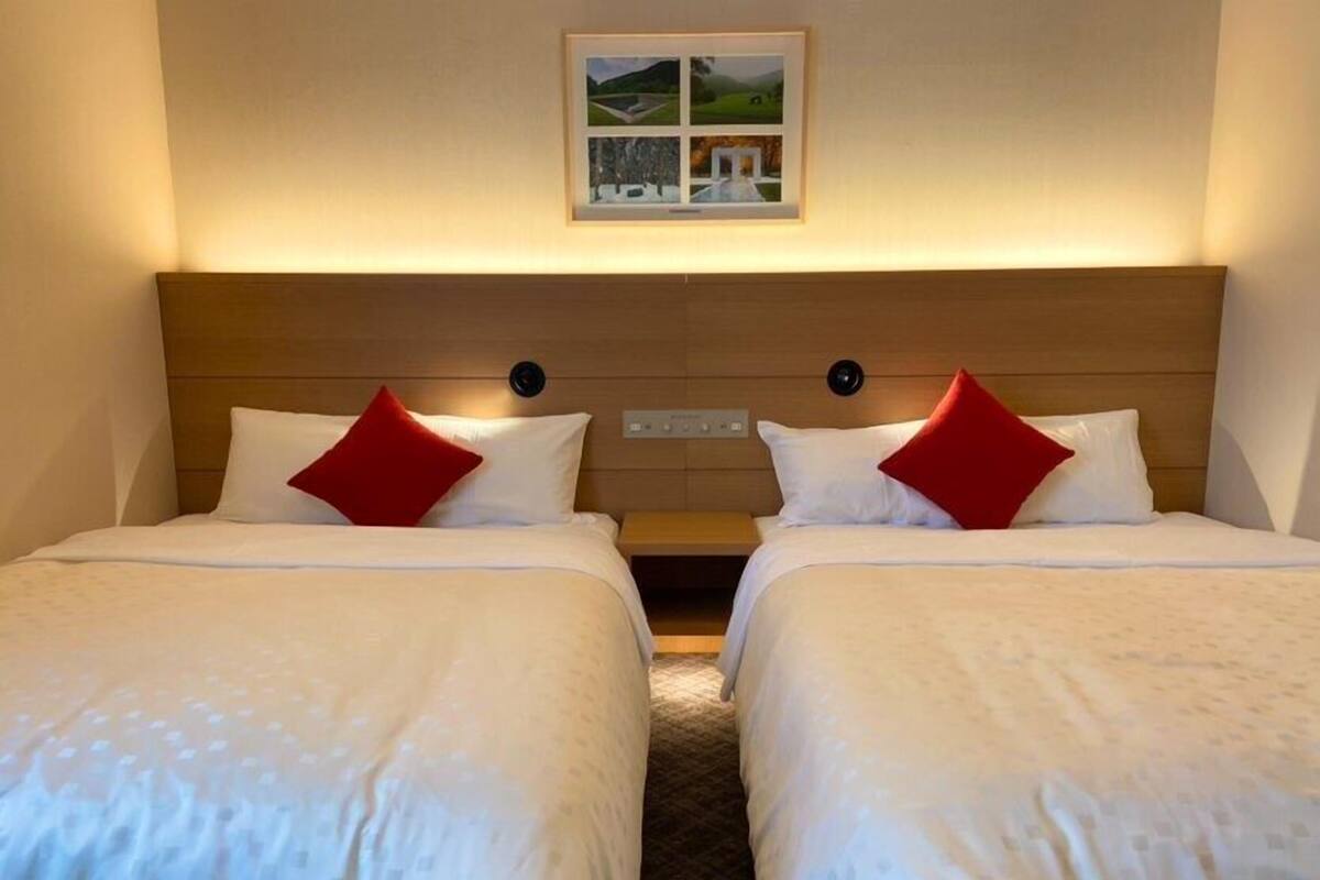 A hotel room with two beds, each with white bedding and red pillows. There is a framed picture above the beds, and a wooden headboard.