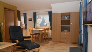 Ein Wohnzimmer mit einem schwarzen Ledersessel, einem Holzespeisetisch mit Stühlen, einer Holzkommode mit Büchern und einem Fernseher an der Wand.