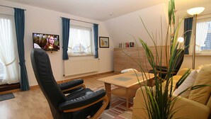 Ein Wohnzimmer mit einer Couch, einem Sessel, einem Couchtisch, einem Fernseher und einem Fenster mit Vorhängen.