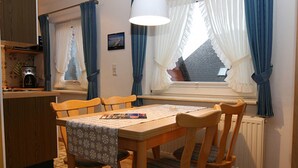 Ein Essbereich mit Holztisch und Stühlen, ein Fenster mit Vorhängen, ein Heizkörper und eine Küchenzeile mit einer Kaffeemaschine.