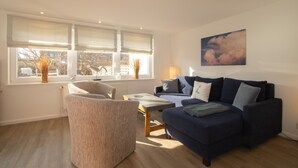 Een woonkamer met een bank, fauteuil en een tafel. Er zijn ramen met zonwering, een radiator en een schilderij aan de muur.