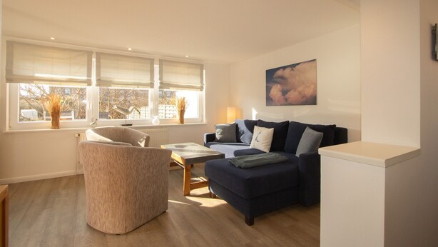 Een woonkamer met een bank, fauteuil en een tafel. Er zijn ramen met zonwering, een schilderij aan de muur en een radiator.