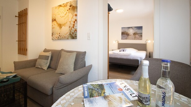 Una sala de estar con un sofá, una mesa con revistas y botellas, una cama al fondo y una pared con un cuadro grande.