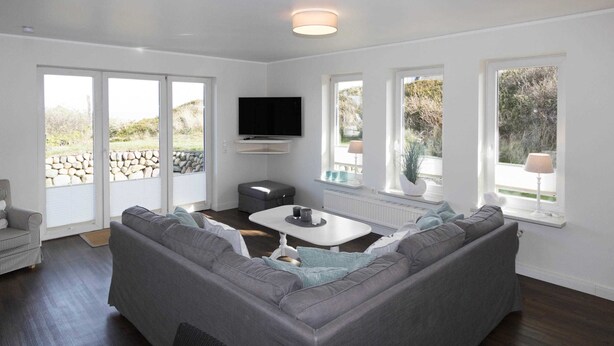 Un salon moderne avec un canapé d’angle gris, une table basse blanche et un téléviseur à écran plat fixé au mur. De grandes fenêtres laissent entrer la lumière naturelle et offrent une vue sur l’extérieur.