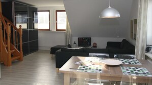 A modern living room with a wooden staircase, a black sofa, a dining table with a placemat, and a pendant light. - House Schössler apartment 2 (Hörnum (Sylt))