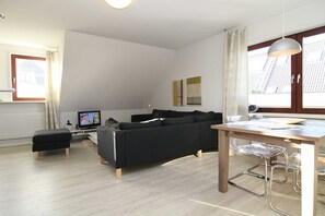 Een moderne woonkamer met een zwarte bank, een houten eettafel en een transparante glazen barstoel.