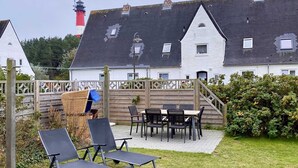 Een terras met buitenmeubilair, een houten omheining en uitzicht op een wit gebouw met een rood-wit lantaarnhuis op de achtergrond.