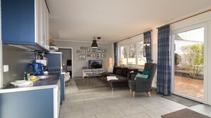 Een moderne keuken met een ingebouwde kookplaat, een koffieapparaat en een spoelbak. Er is een zithoek met een bank en een televisie. De kamer heeft grote ramen met gordijnen en uitzicht op buiten.