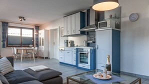 Een moderne keuken met witte en blauwe kasten, een magnetron en een fornuis. Er is een eethoek met een tafel en stoelen, en een bank in de woonkamer. Een glazen salontafel bevat een fles champagne en een bord met taart.