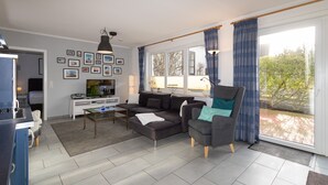 Een moderne woonkamer met een bank, fauteuil en salontafel. Er hangen ingelijste foto’s aan de muur, er is een televisie en in de achtergrond is een keuken te zien.