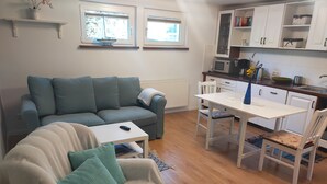 Ein Wohnzimmer mit einer Couch, einem Couchtisch und einem Esstisch mit Stühlen. Zwei Fenster mit Jalousien, eine Mikrowelle und eine Schrank mit Regalen sind ebenfalls vorhanden.
