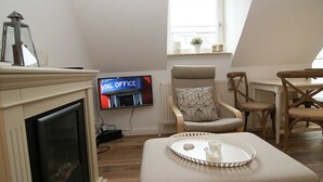 Un salon chaleureux avec une cheminée, un téléviseur diffusant l'émission « WILL OFFICE », et un coin repas avec des meubles en bois.