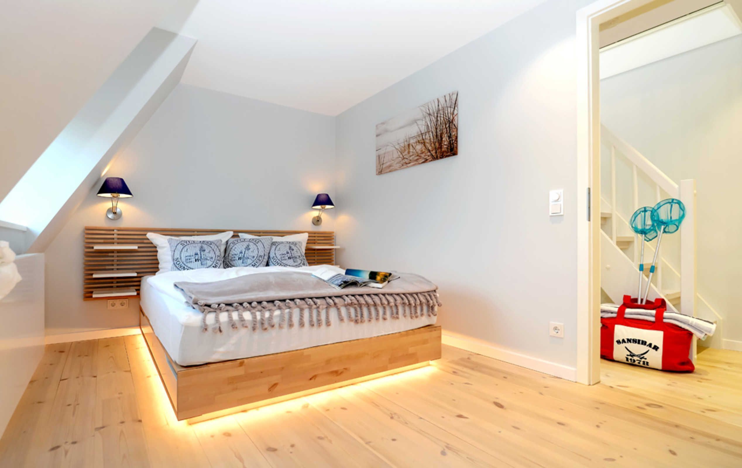 A modern bedroom with a sloped ceiling, a wooden bed frame, two bedside lamps, a wall-mounted artwork, and a red bag on a wooden table.