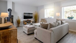 A modern living room with a sofa, ottoman, coffee table, and a flat-screen TV mounted on the wall. There are two windows providing natural light, and a small table with a lamp and decorative items. - Sylt Hüüs, Germany (Hörnum (Sylt))