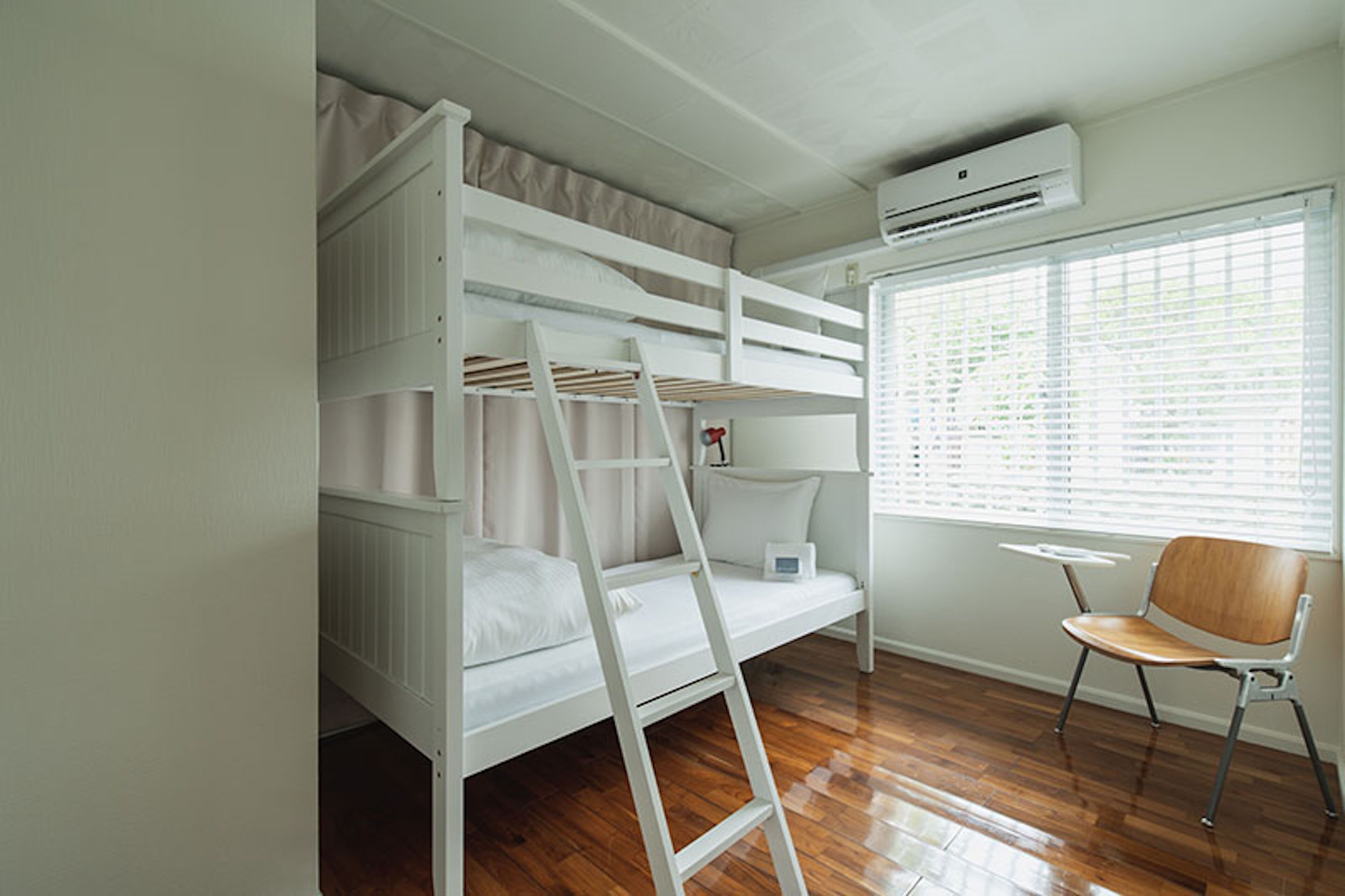 Una cama litera con escalera, una mesita y una silla en una habitación con suelo de madera y una ventana con persianas.