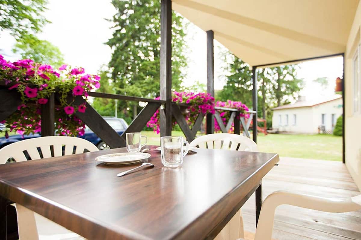 A covered patio with a wooden table, white chairs, and pink flowers.