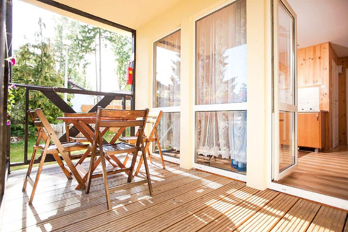 A wooden deck with a table and chairs, large sliding glass doors, and a view of greenery outside.