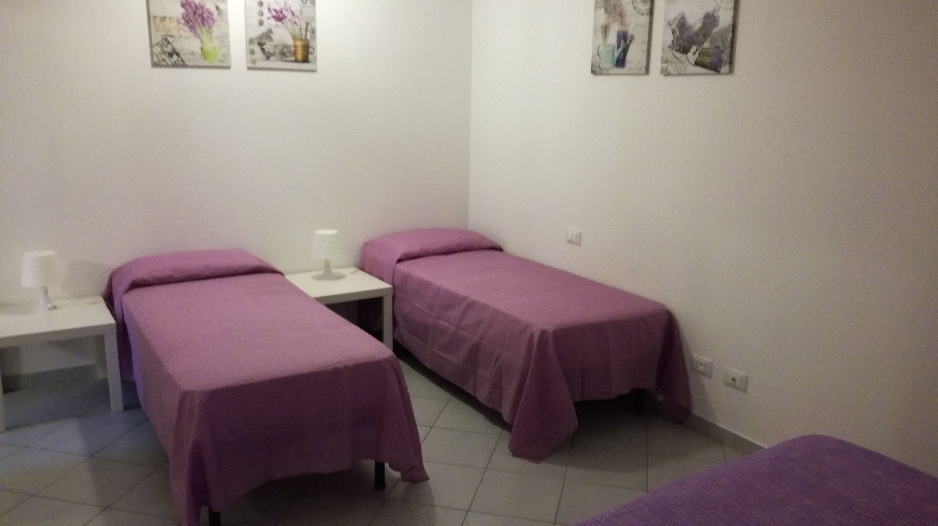 A room with two single beds, each with a purple bedspread, a bedside table with a lamp, and a wall-mounted artwork above the beds.